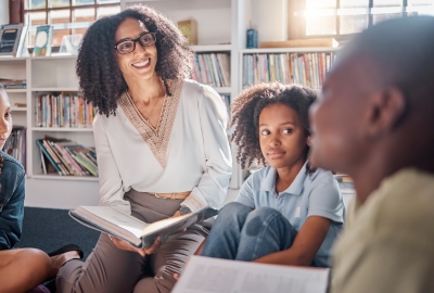 The image shows a woman with curly hair and glasses wearing a white blouse with intricate embroidery at the neckline. She is sitting and holding an open book. Around her are three children: one with braided hair, one with curly hair, and one with short hair. They are seated on the floor in a library. Behind them are shelves filled with books, and sunlight is coming through a window in the background.