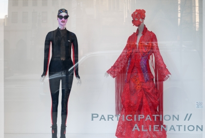 A close-up of a fashion exhibition curated by Costume Studies students. Two dress forms, one in a tight-fitting black suit, and one in a flowing red dress and a silly hat.