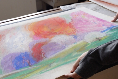 Two hands hold a colorful pastel drawing of a landscape