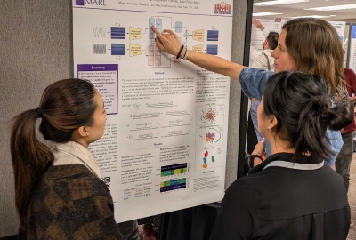 Two members of MARL point to their poster presentation, explaining the findings to a conference attendee. The project they are presenting is "Self-Supervised Mult-View Learning for Disentangled Music Audio Representations"