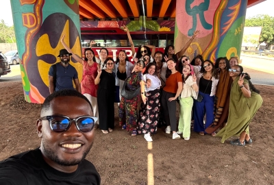 A smiling man in sunglasses takes a selfie while a group of students poses in the background under a brightly-painted bridge