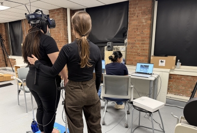 Participant stands on balance board while wearing VR headset, while one research assistant supports them with a hand on their back, and another research assistant operates a computer nearby