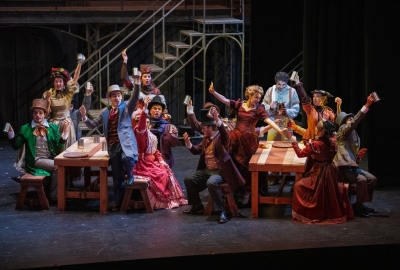 Actors on stage sitting at two long tables singing and toasting each other