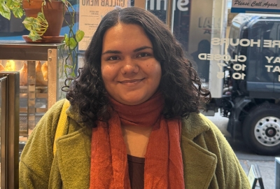 Photo of Laura Assanmal standing in a shop, wearing an orange scarf and green jacket