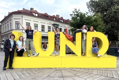 Students pose on and around a bright yellow sign reading "Lund"