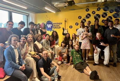 A group of students sits in front of a yellow wall that reads "Warner Music Group"