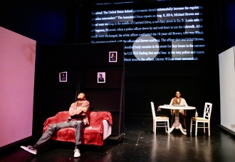 Two actors sitting on stage. one is a red sofa and the other at a table