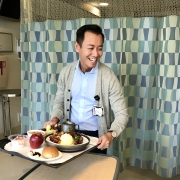 Andy Chu holding a tray of hospital food in front of a medical curtain with a geometric pattern on it.