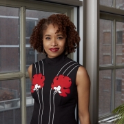 Dr. Nicole R. Fleetwood stands next to a window wearing a black dress with white stripes and red flowers.