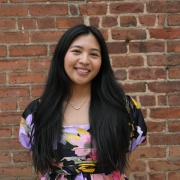 A portrait of a smiling Kira Santos, T&L Alumni, in front a brick wall and wearing a floral dress.