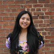 A portrait of Kira Santos in a floral dress, standing in front of a brick wall