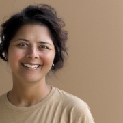 A woman with tan skin, short black hair, a tan shirt, is smiling. 