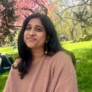 A portrait of Vaishnavi Akilla wearing a coral colored sweater in a park with beautiful trees in the background