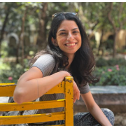 The image captures a photo of PRE researcher Rhea Almeida. She smiles in the photo, while turning back in chair to engage the camera. Rhea sits in a wooden chair placed in a green space.