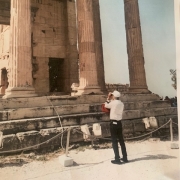 Gerald standing in front of an ancient greek temple taking a photo. He's wearing a white long sleeve shirt, black pants, and a white cap.