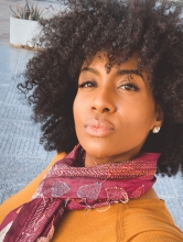 Brown woman with curly hair, purple scarf, and mustard-gold sweater posing for a selfie