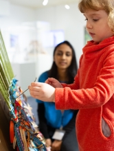 An Art + Education MA student observes a young child in a classroom. The child is in the foreground wearing a red shirt and touching a loom. The child is focused on their task. The teacher sits in the background and is slightly out of focus. The teacher has long dark hair and wears a blue sweater.