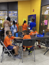 Children in orange shirts sit around a table reading and talking. In the back, families line up to receive a book from a vending machine. 