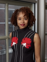 Dr. Nicole R. Fleetwood stands next to a window wearing a black dress with white stripes and red flowers.