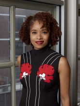 Dr. Nicole R. Fleetwood stands next to a window wearing a black dress with white stripes and red flowers.