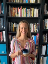 Rachel Northrop, holding a mug, smiles in front of a bookshelf full of books. 