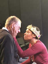 A Black woman, wearing a gold crown, cradles the face of an older white man as they face each other. 