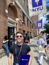 Dylan Licastro, wearing an NYU Steinhardt lanyard and sunglasses, smiles in front of an NYU building. 