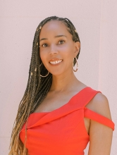 Photo of Heather Clarke a Brown Skinned Black monoracial Autistic woman with long waist length braids wearing a read top and gold hoop earrings. She is looking ahead at photo and smiling. 
