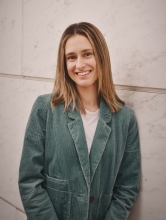 A woman in a teal blazer with shoulder length blond hair leans against a wall and smiles at the camera