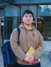 The photo depicts a professional photo of Mars Jiang in a brown sweater holding a yellow book and blue and black bookbag outside