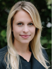 Headshot image of Elle, she has blond hair and is wearing a navy coat. She is smiling center frame and standing in front of greenery which is out of focus.