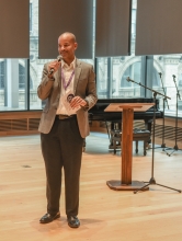 Jonathan Jones standing, speaking into a mic at the anniversary event. Behind him sites a panelist, listening.