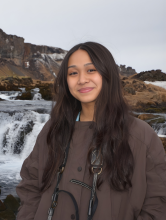 A young woman with long dar brown hair parted in the middle stands in front of a waterfall. She has a camera strap around her neck and is wearing a dark brown jacket. 