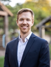 Portrait of Andrew E. Koepp smiling outdoors, wearing a navy blazer and light blue button-down shirt, standing in front of a softly blurred natural setting with rustic wooden structures in the background.