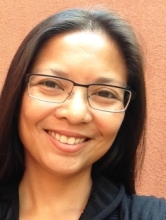Portrait of Lara Saguisag smiling against a warm peach-colored wall, wearing rectangular glasses and a black top, with long dark hair worn down.