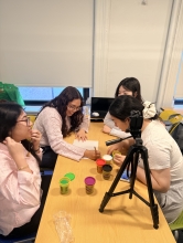 A photo of 4 people interacting around a table with play dough, a camera stand, and one person smiling, writing on a piece of paper
