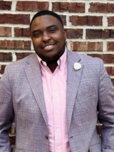 Portrait of Gabriel Lockett smiling in front of a red brick wall, wearing a light gray blazer over a pink button-down shirt with a white lapel pin.