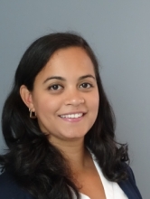 Portrait of Lillian Polanco-Roman smiling against a gray studio background, with shoulder-length dark hair, wearing a navy blazer over a white blouse and small hoop earrings.