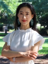 Portrait of Shuang Fu standing outdoors in a park-like setting, smiling with her arms crossed, wearing a white blouse with ruffled sleeves and turquoise statement earrings against a sunlit green background.