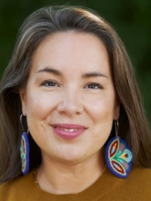 Portrait of Eve Tuck smiling outdoors, with long dark hair, colorful beaded earrings, and a mustard-yellow top against a softly blurred green background.