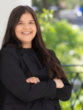 Portrait of Sophia Rodriguez smiling outdoors with her arms crossed, wearing a black blazer and black top, standing in front of a softly blurred green and architectural background.