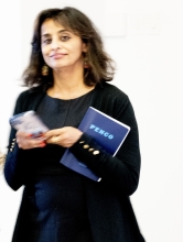 A South Asisan woman with shoulder long chestnut hair holds a book and a phone and looks at the camera.