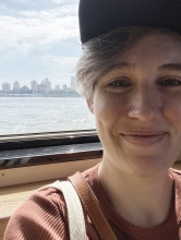A white woman in her 30s with short hair, in front of a window with a skyline view.