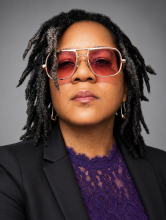 African-American woman with rose tinted glasses and locs wearing a black blazer and violet lace top