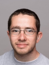 Visual Description  Subject: A young man with short brown hair, wearing metal-rimmed eyeglasses and a light gray crewneck shirt.  Expression: A subtle, gentle smile with a neutral gaze directed forward.  Background: A solid, plain light gray studio backdrop.