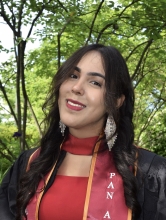 A young woman is shown in front of some trees with long dark hair, large dangling earrings, and has a graduation sash around her neck.
