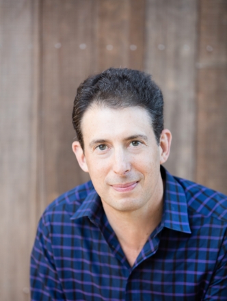 portrait of Eric Klinenberg wearing a blue plaid shirt  in front of a wood wall