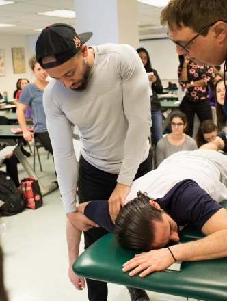 A man, laying facedown on a padded medical table, has his arm manipulated by a physical therapy student while an instructor and classmates look on.