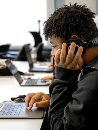 A student works on his laptop, holding headphones to his ears