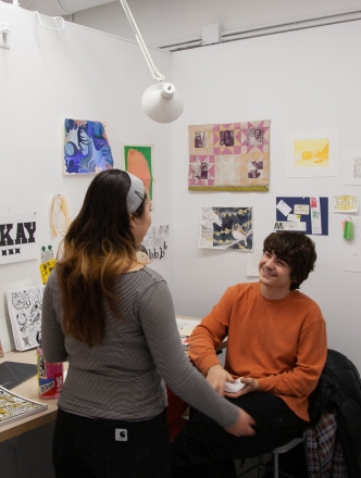 Two students talking in a BFA studio, colorful artwork is pinned on the wall behind them